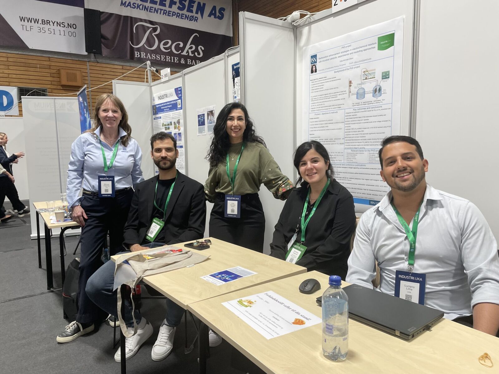 Fem deltakere poserer ved et bord på en messestand med plakater og skillevegger i bakgrunnen.