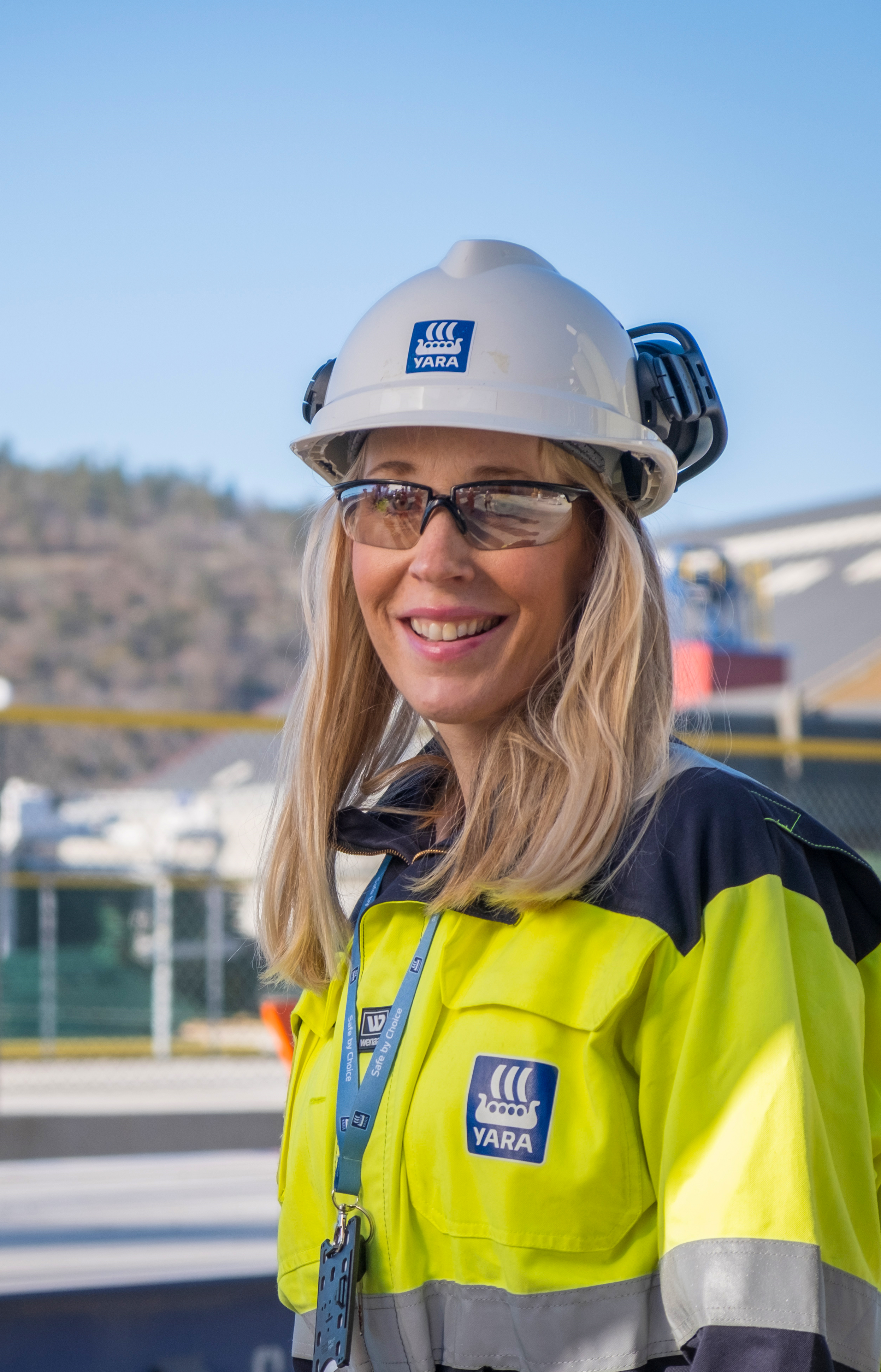 Smilende person med hjelm, vernebriller og gul synlighetsjakke foran et industriområde utendørs.