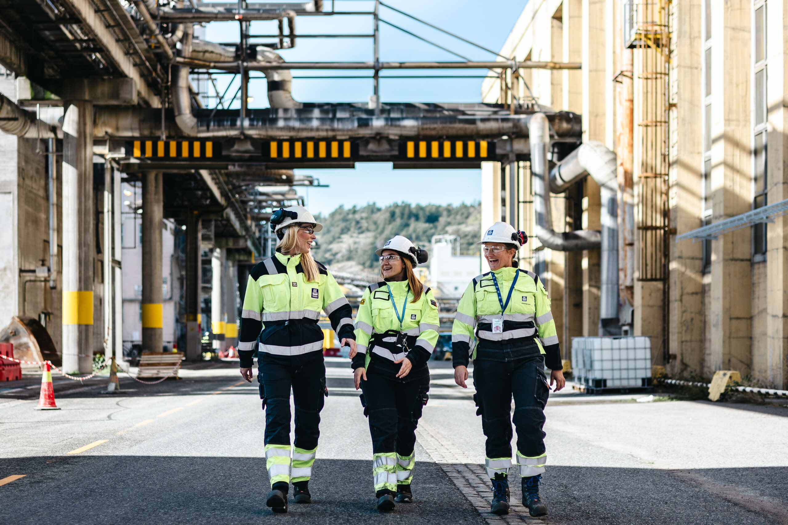 Tre personer i verneklær og hjelmer går sammen mellom industribygg og rørføringer.