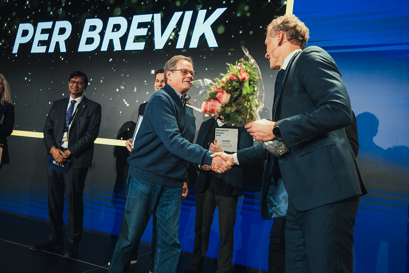 To menn håndhilser på en scene mens den ene holder blomster og diplom, med navnet Per Brevik på skjermen bak.