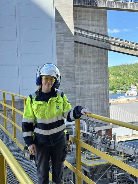 Person med hjelm, hørselsvern og synlig arbeidstøy står ved et gult rekkverk på et industriområde.