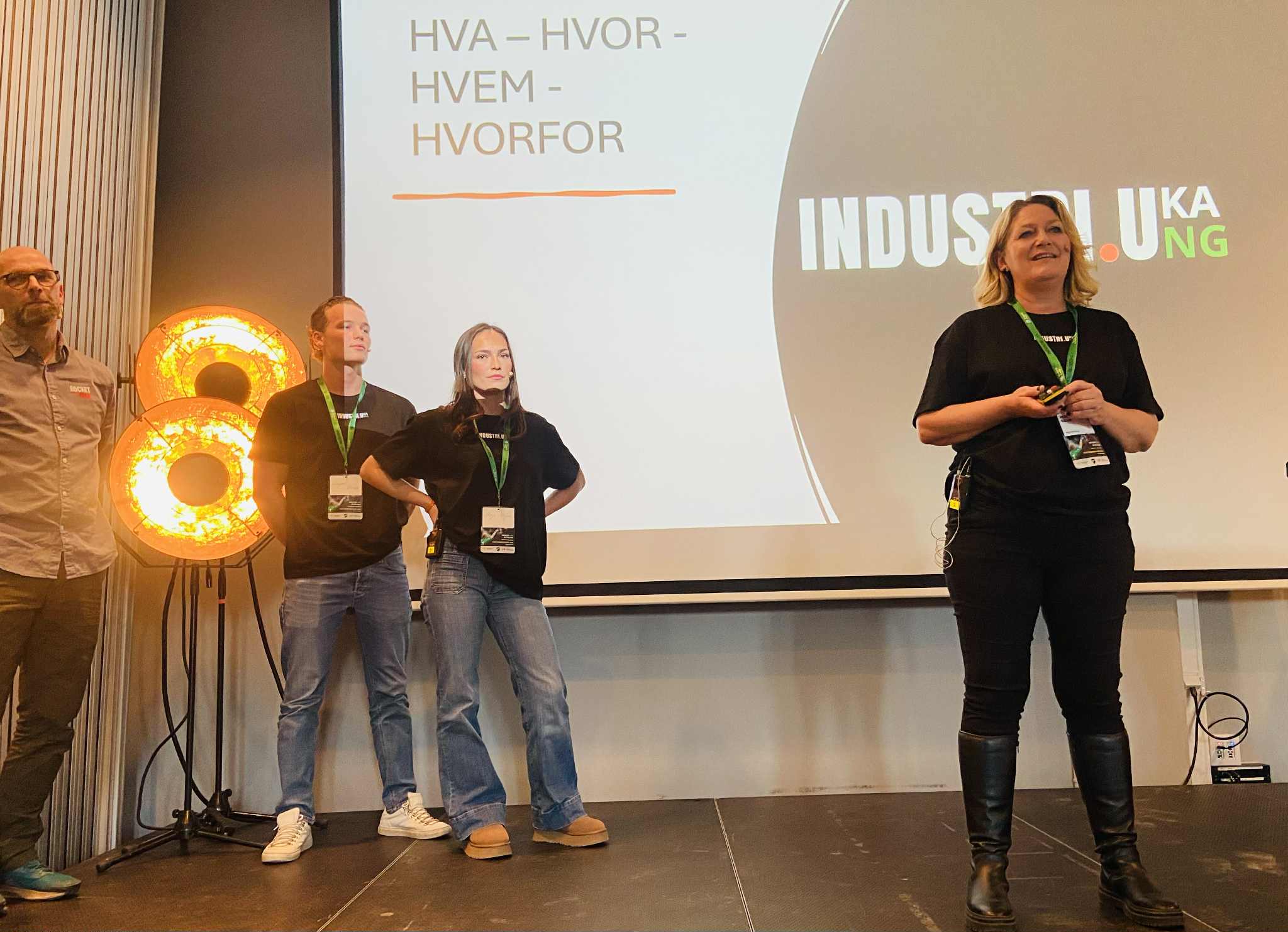 Fire personer står på en scene foran en presentasjon, mens en kvinne til høyre holder fjernkontroll og snakker.