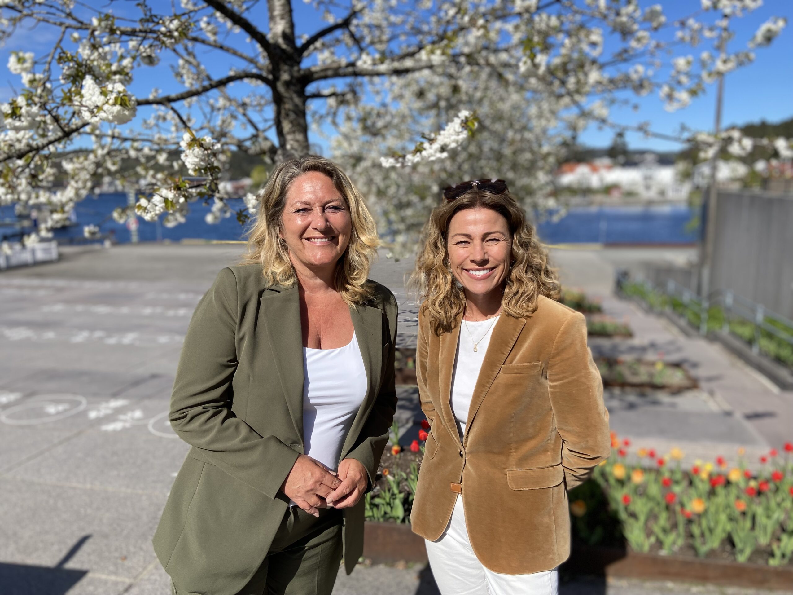 To smilende kvinner står utendørs foran et blomstrende tre, med vann, bygninger og tulipaner i bakgrunnen.