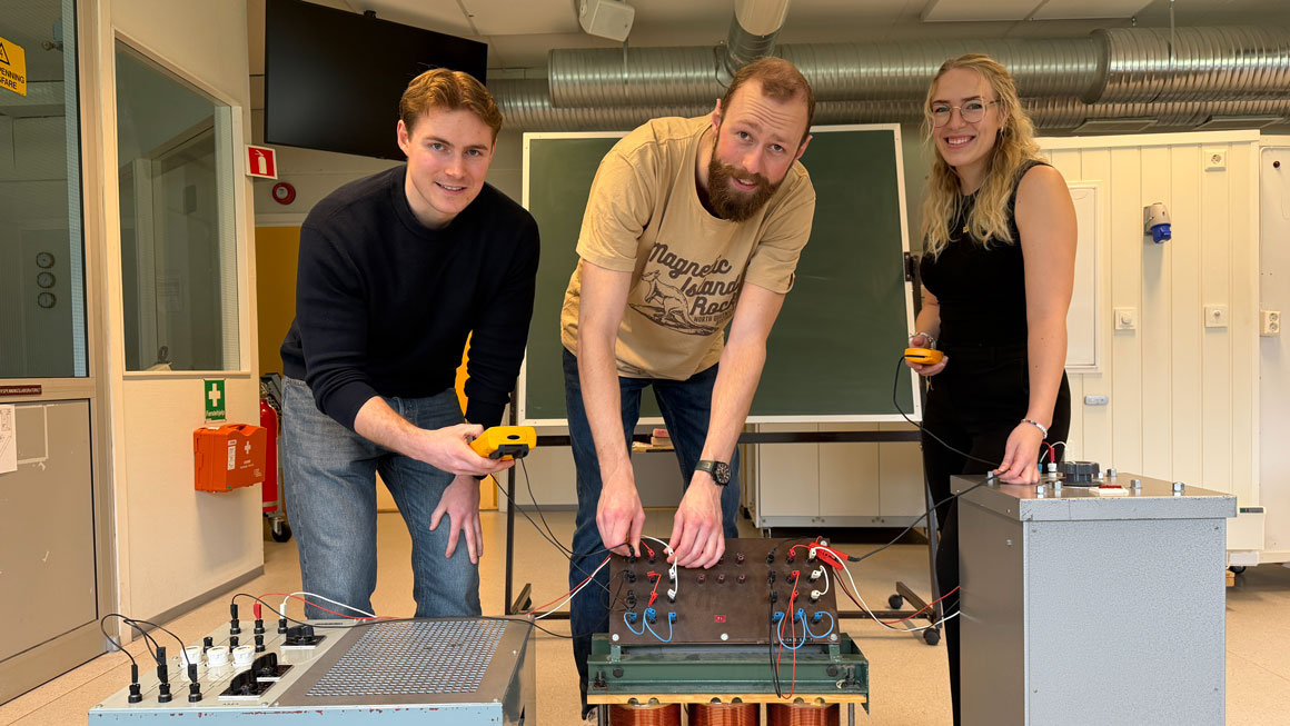 Tre personer står ved elektrisk laboratorieutstyr med ledninger og måleinstrumenter i et undervisningsrom.