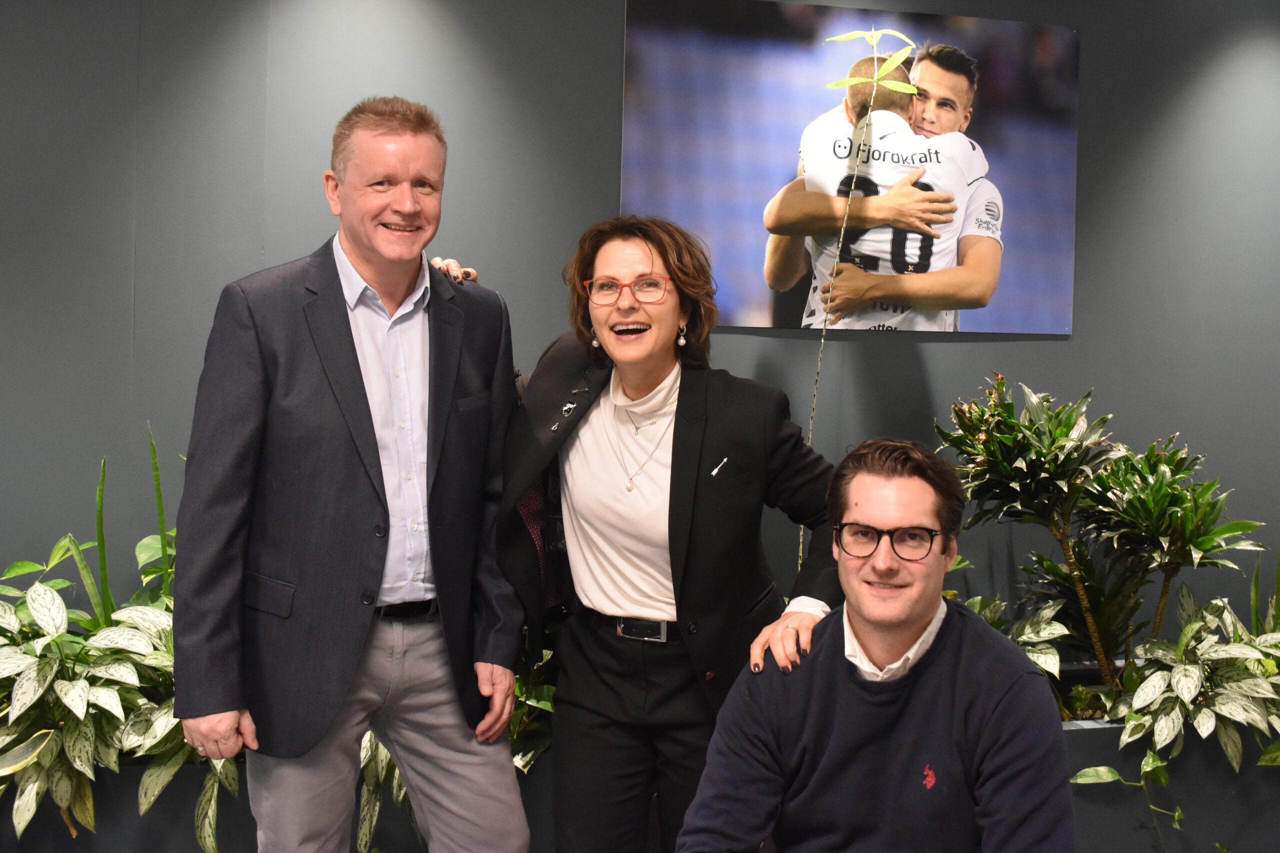Tre personer poserer foran en vegg med planter og en skjerm som viser fotballspillere i omfavnelse.