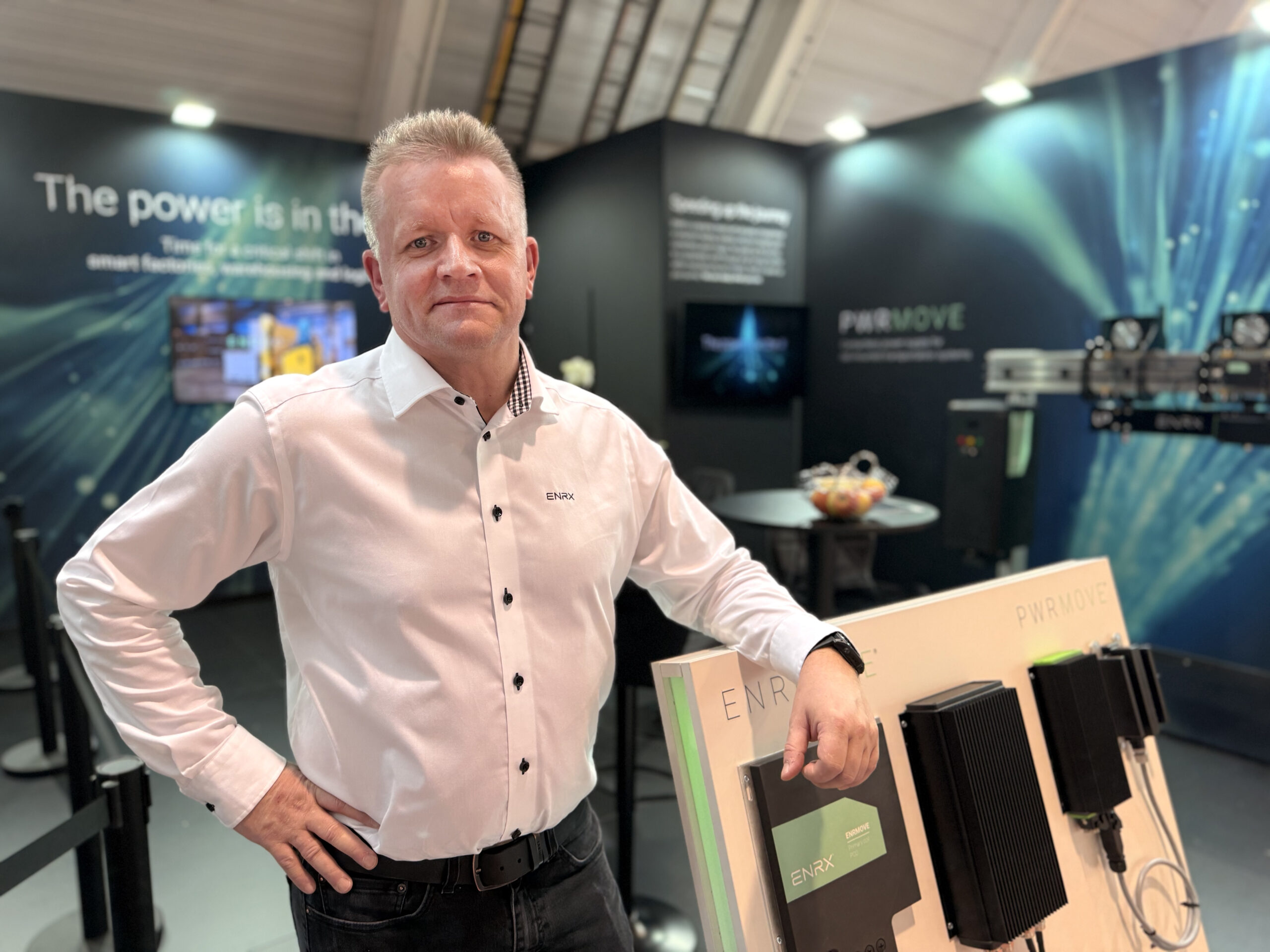 Mann i hvit skjorte står ved en utstillingsstand med elektronisk utstyr og skjermer i bakgrunnen.