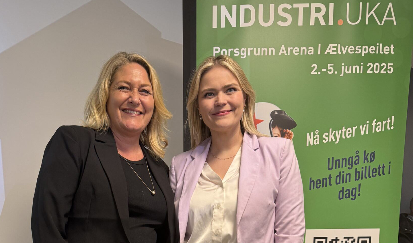 To smilende kvinner står foran en grønn plakat for Industriuka med informasjon om arena og dato.