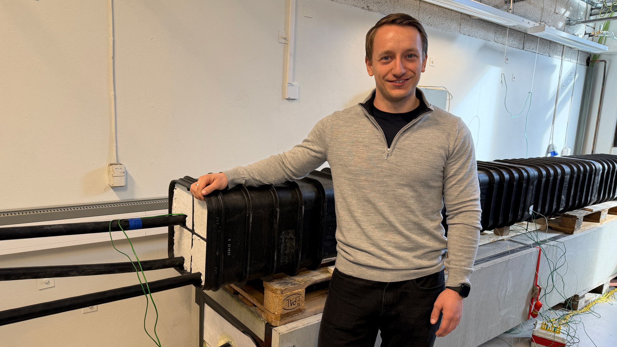 Mann står ved en lang svart kabel- eller rørmodul på paller i et teknisk rom med ledninger og måleutstyr.